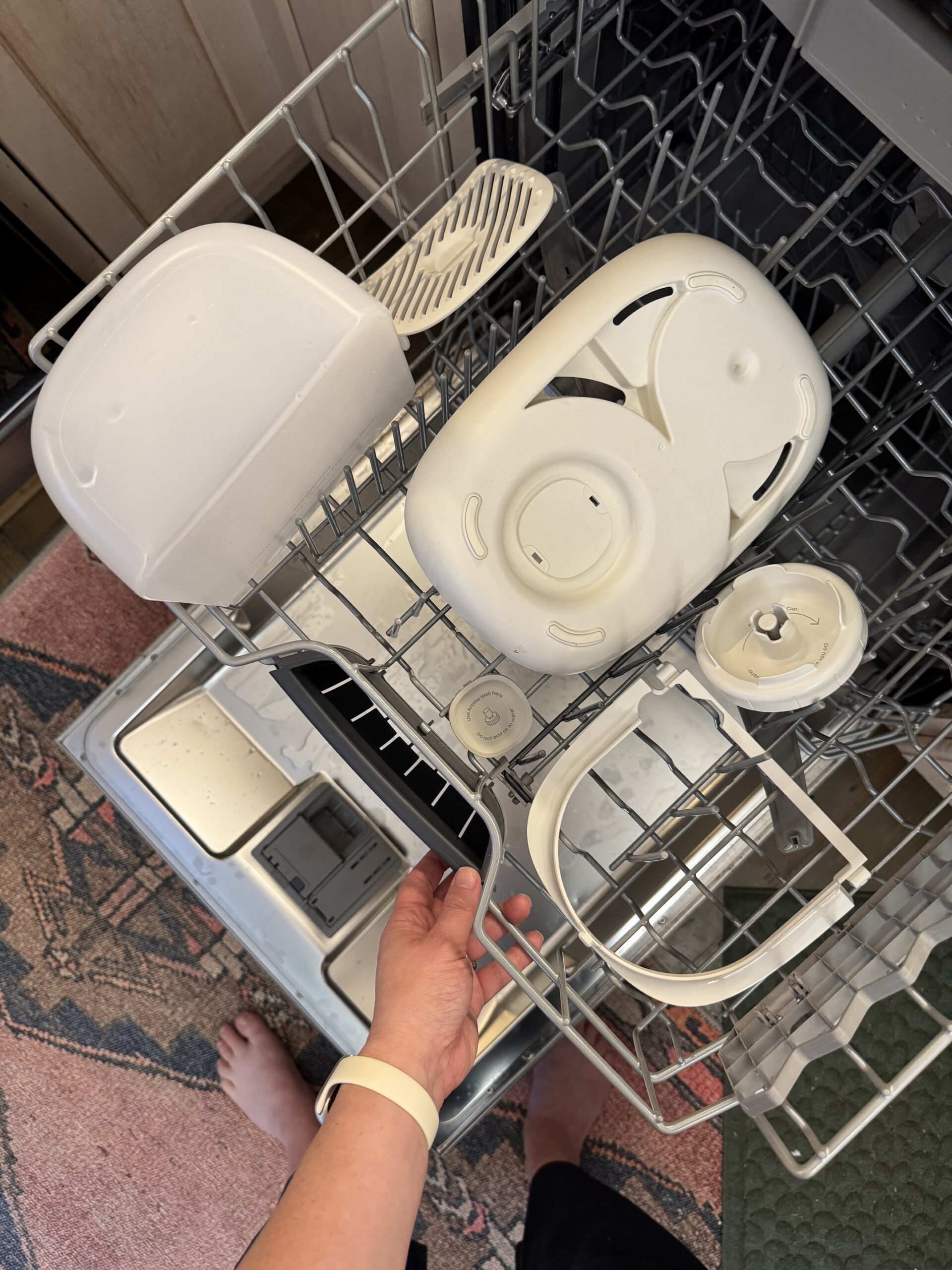 A person loads white plastic parts, likely from a kitchen appliance, into the top rack of a dishwasher. The dishwasher door is open, and a patterned rug is visible on the floor.