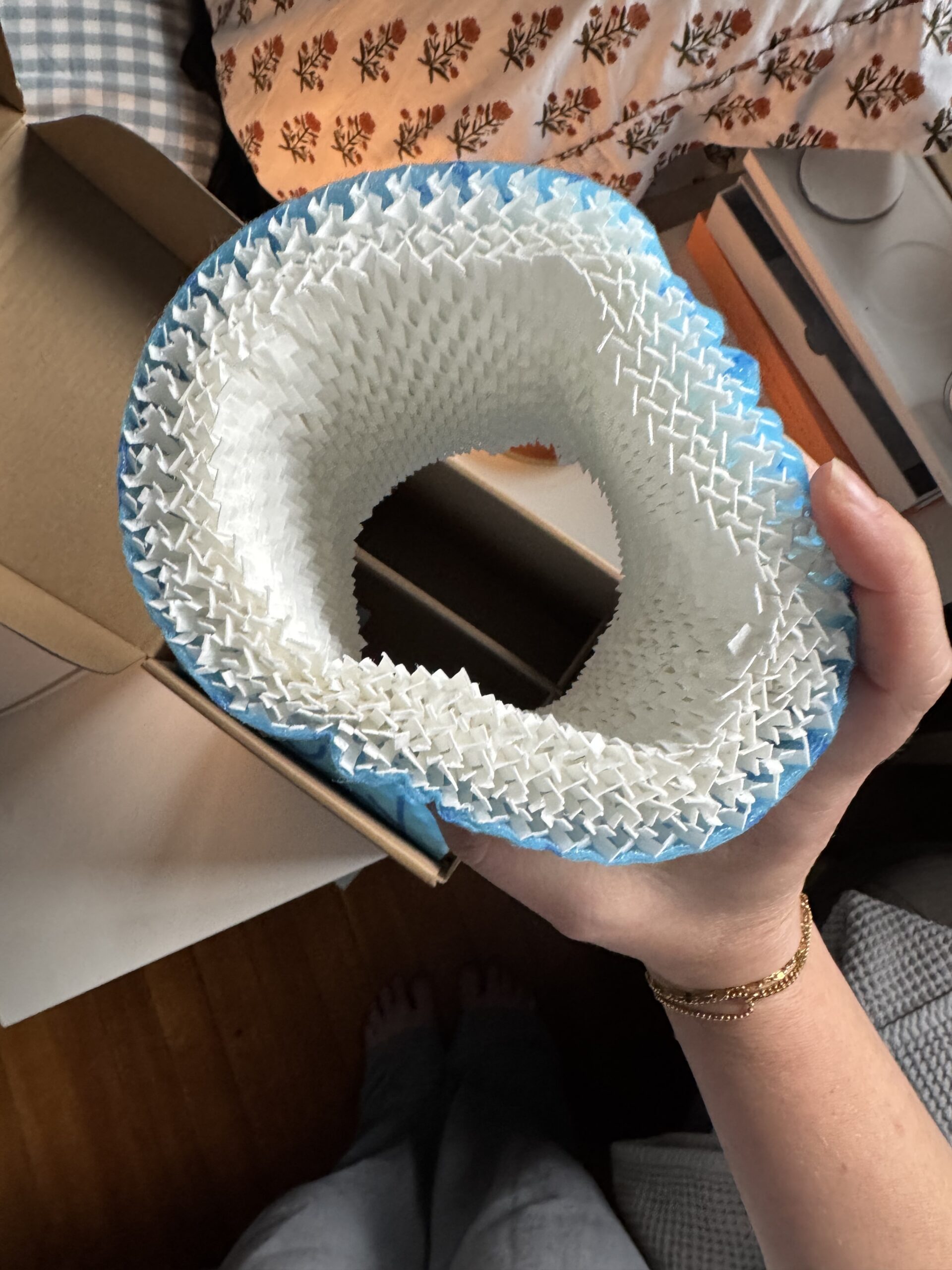 A hand holds a white and blue flexible mesh cylinder, possibly packaging material, over a wooden floor and cardboard box.
