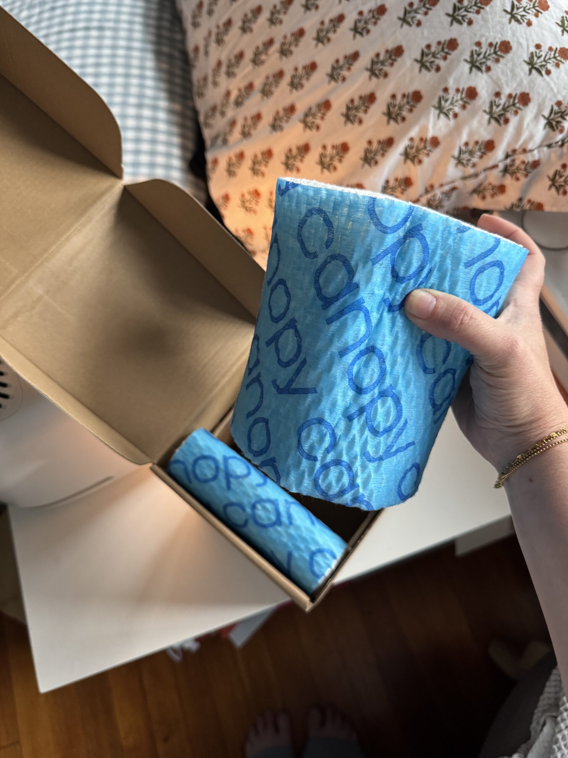 A hand holds a blue patterned paper roll labeled "canopy" above an open box containing another identical roll, near a bed with floral sheets.