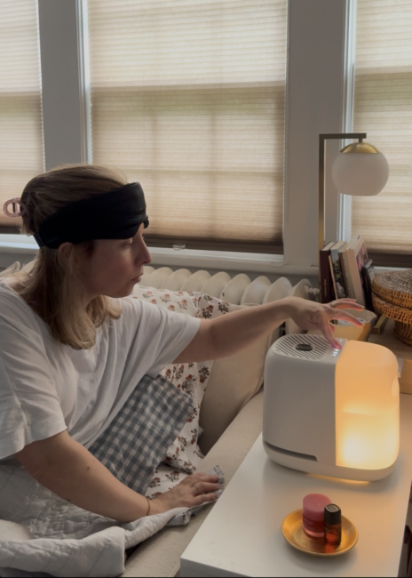 A person wearing a black headband and white shirt sits on a couch and adjusts a white humidifier or diffuser on a side table next to a bed.