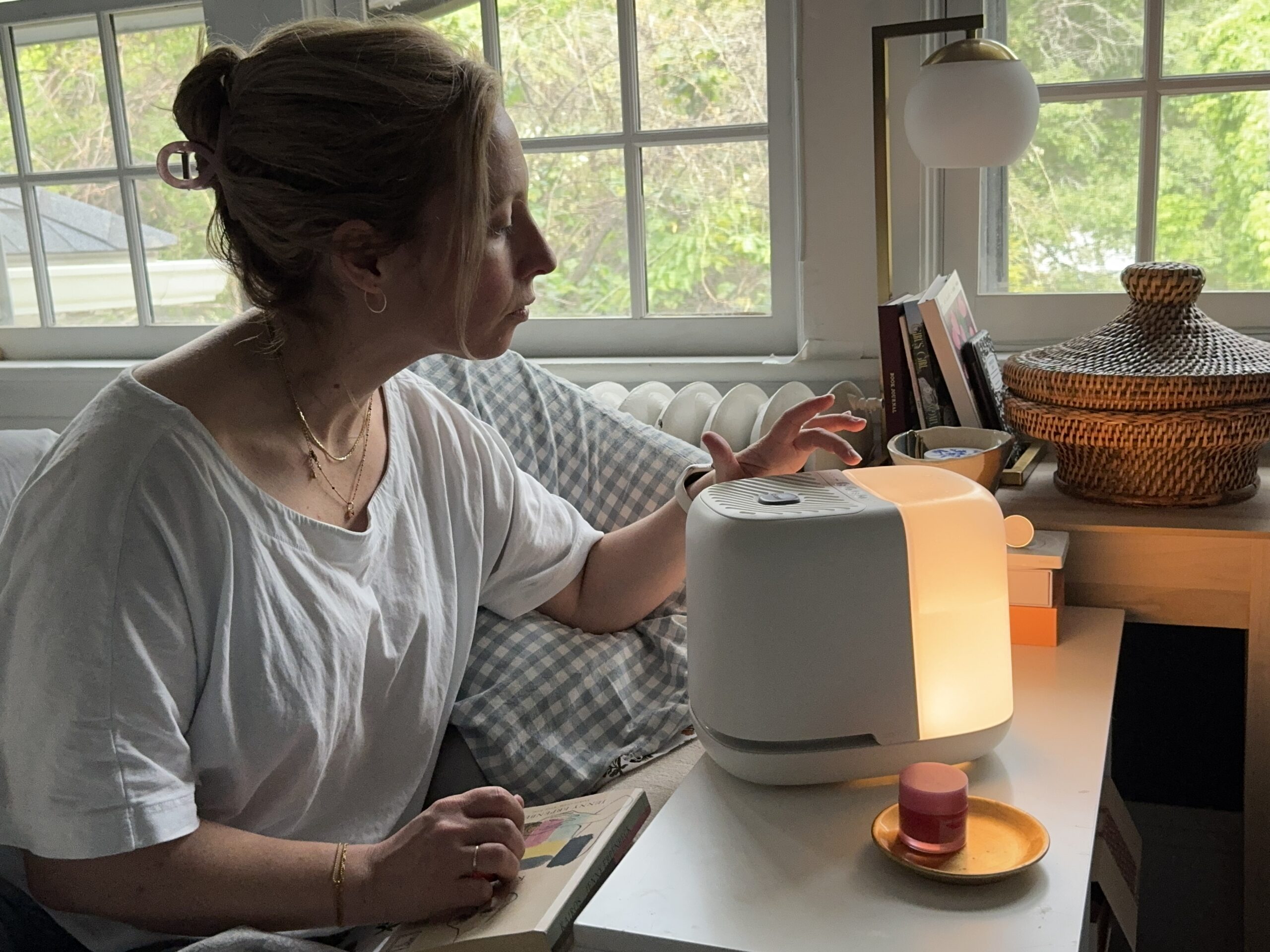 A woman sits on a couch next to a table, reaching toward a white humidifier or diffuser that is lit, with a small container and a book nearby.