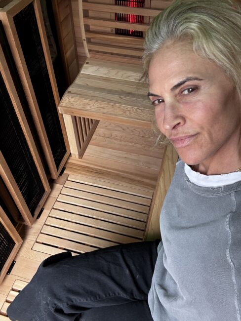 A person with blond hair, wearing a grey top and black pants, sits in a wooden sauna, looking at the camera.