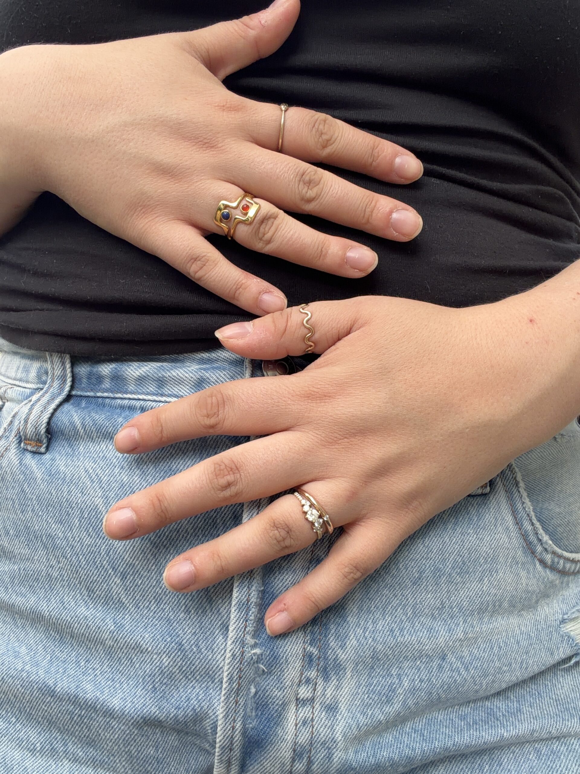 Close-up of a person wearing multiple rings, with hands resting on their stomach and hip, dressed in a black top and light blue jeans.