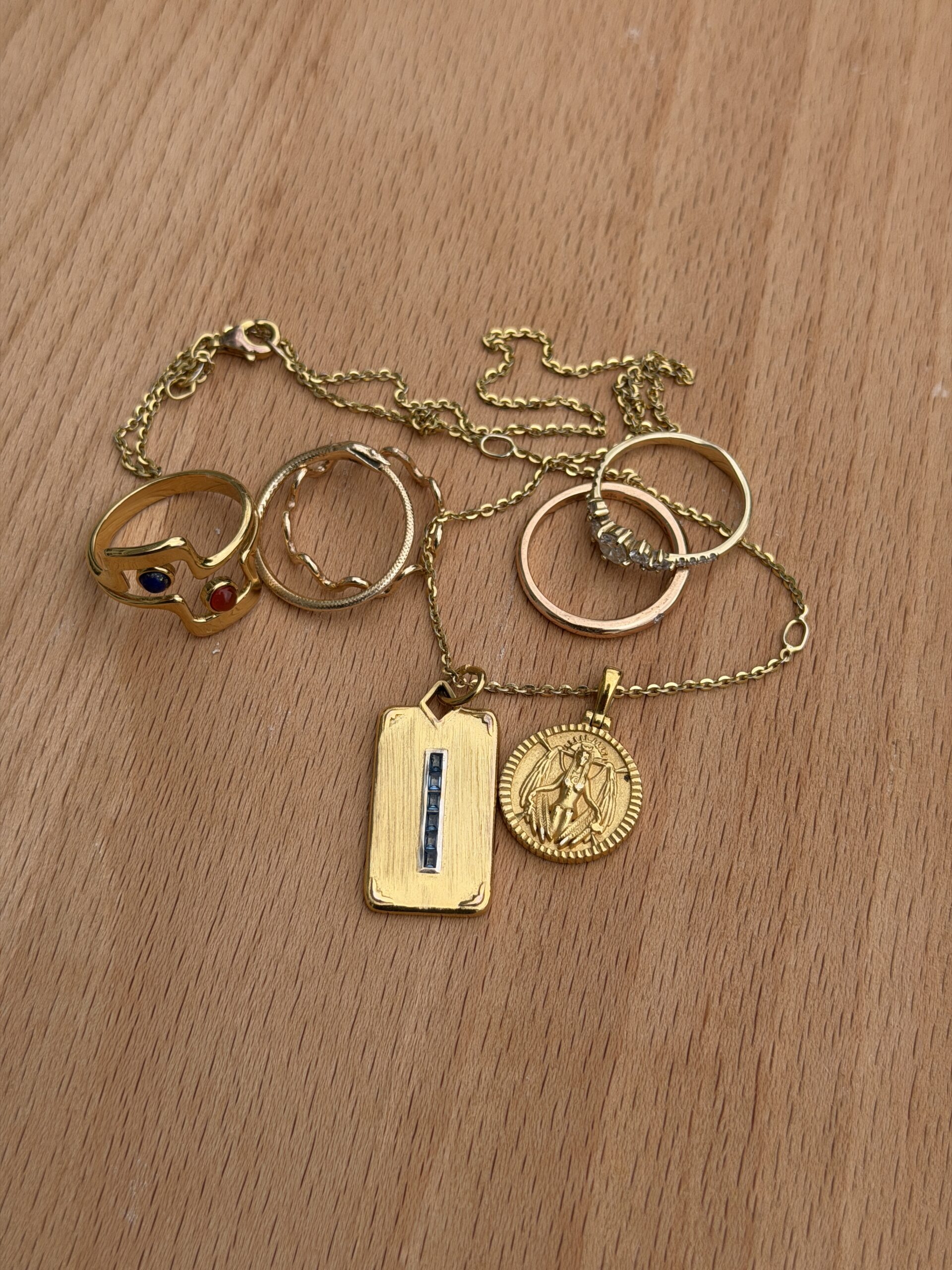 A gold chain with two pendants and four gold rings, one with colored stones, displayed on a wooden surface.