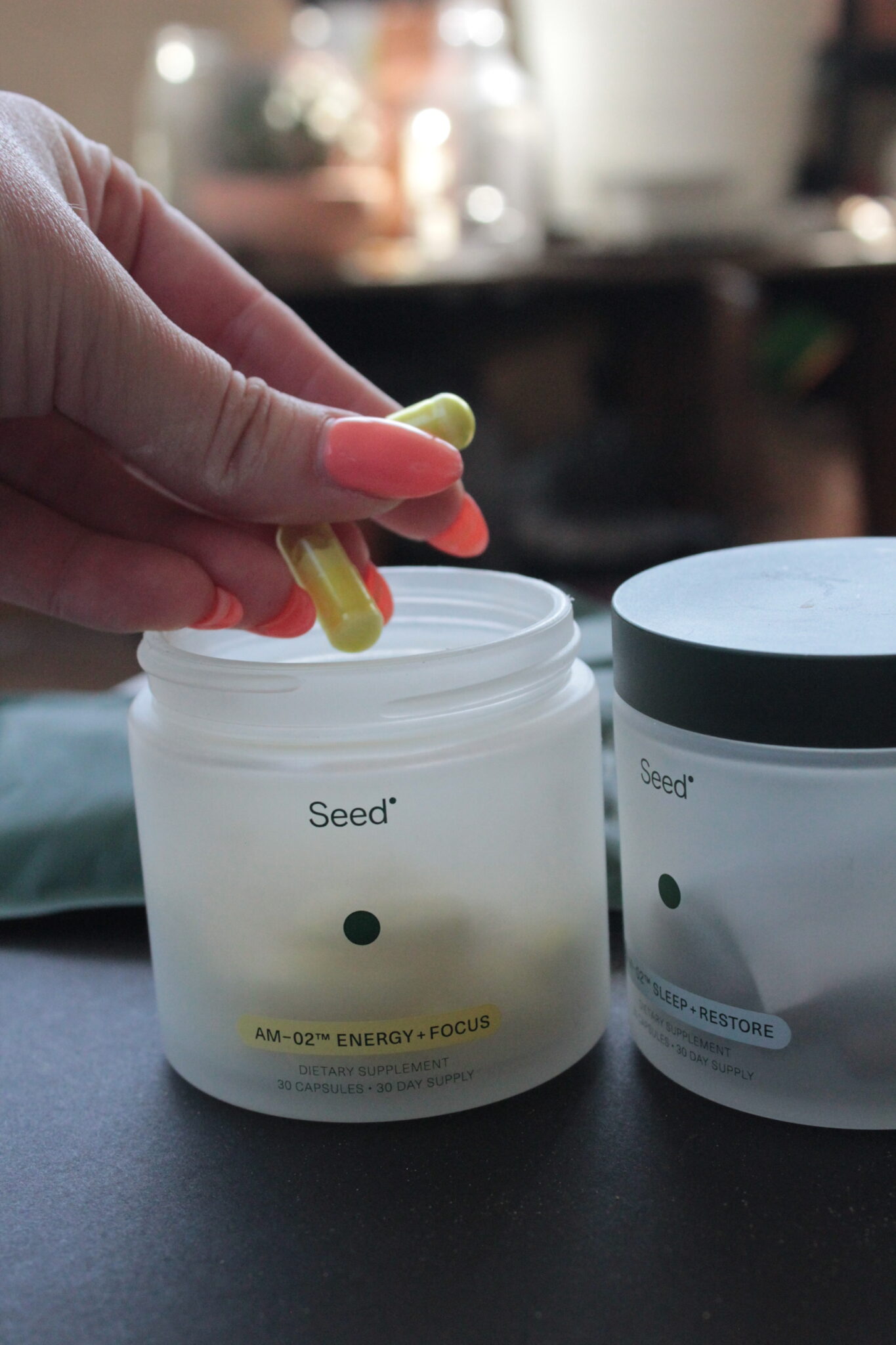 A hand holds two yellow-green capsules above an open Seed supplement container labeled "AM-02™ Energy + Focus." Another labeled container is nearby.