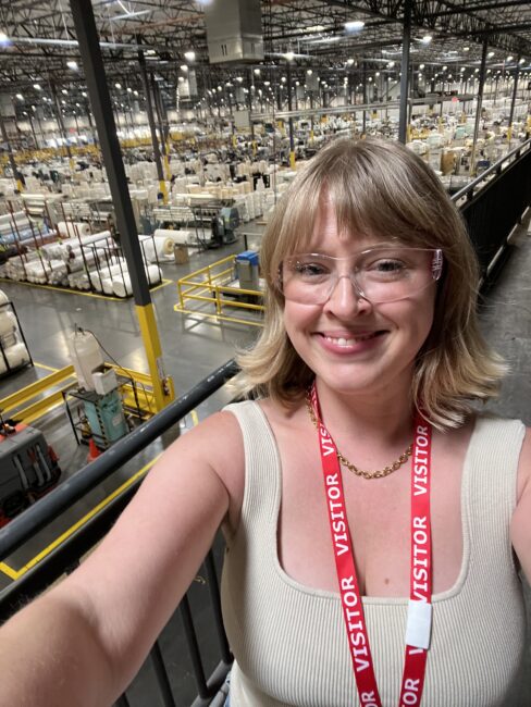 A person wearing a "VISITOR" lanyard takes a selfie in a large warehouse filled with rows of goods and industrial shelving.