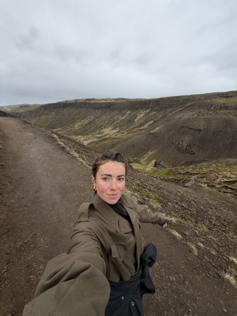 Person taking a selfie on a dirt path in a hilly, rocky landscape under an overcast sky.