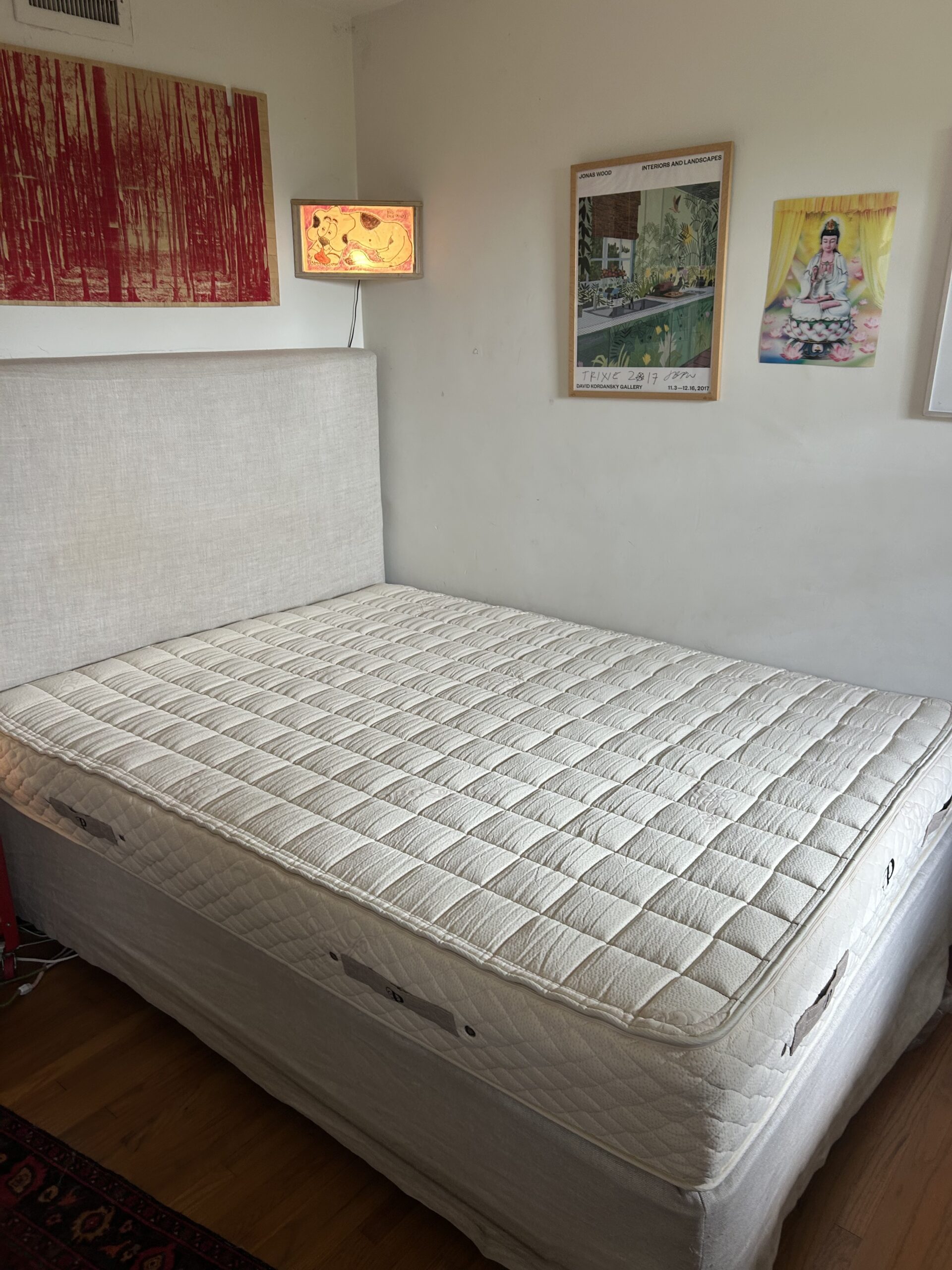 A neatly made beige bed with no sheets or pillows, placed against a white wall with three colorful artworks hanging above it.