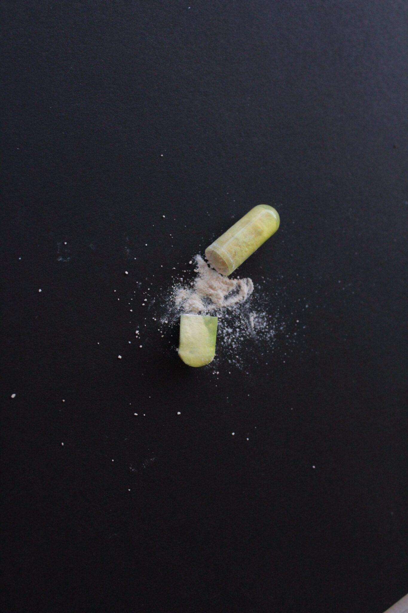 A green capsule pulled apart with white powder spilling out onto a black surface.