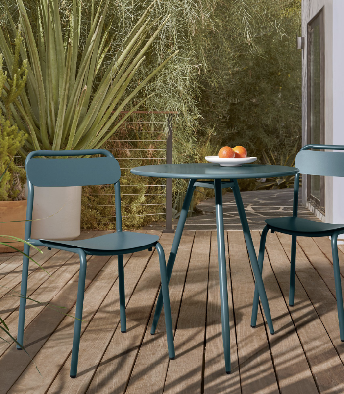 A small round table with two blue chairs on a wooden deck, with a plate of oranges on the table and plants in the background.
