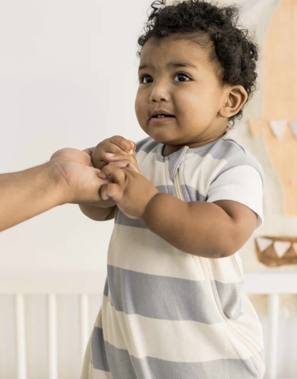 A young child in a short-sleeve, gray and white striped outfit holds onto an adult's hand while standing indoors.