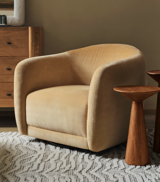 A tan upholstered armchair sits on a patterned rug next to two wooden side tables and a wooden dresser against a beige wall.