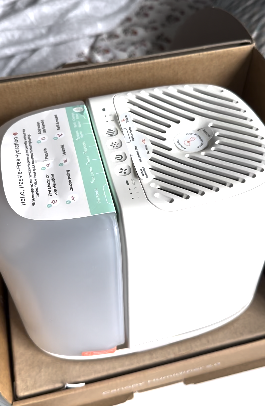 A white countertop humidifier with control buttons and a translucent water tank, placed inside a cardboard box.