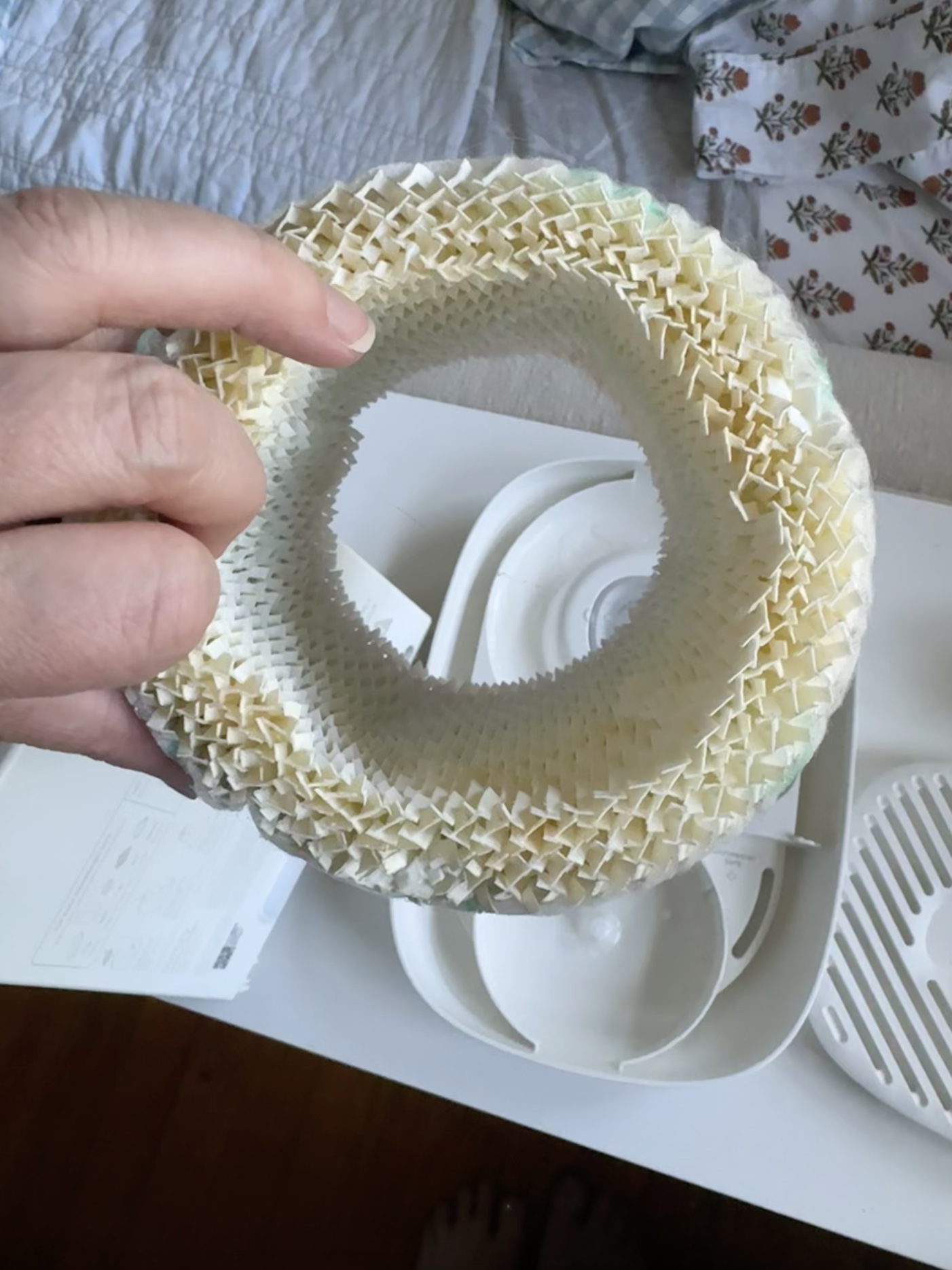 A hand holds a cylindrical, honeycomb-patterned air filter above a disassembled humidifier on a white table.