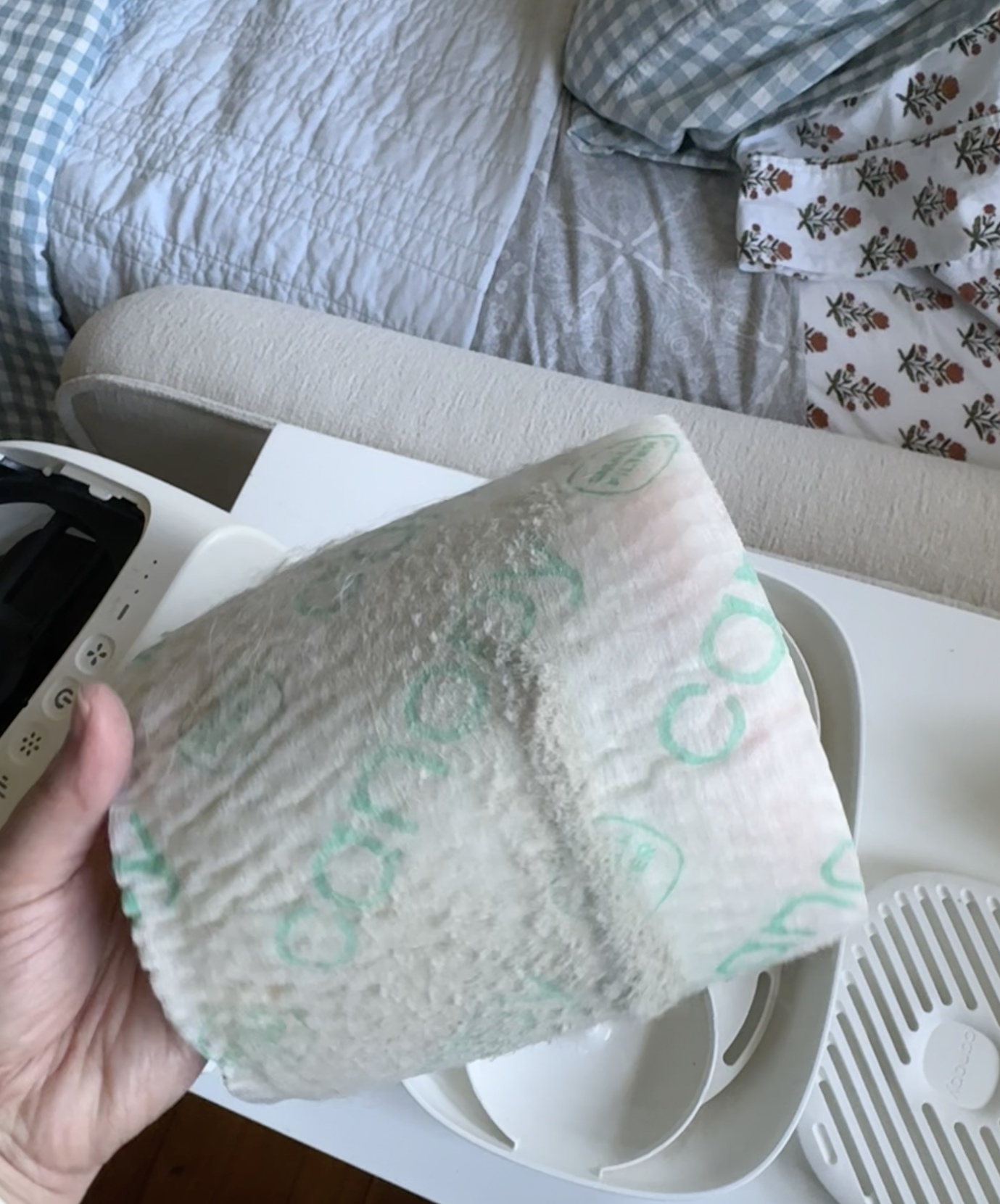 A hand holds a used and dirty air purifier filter above a white table with purifier parts; a bed with patterned sheets is in the background.