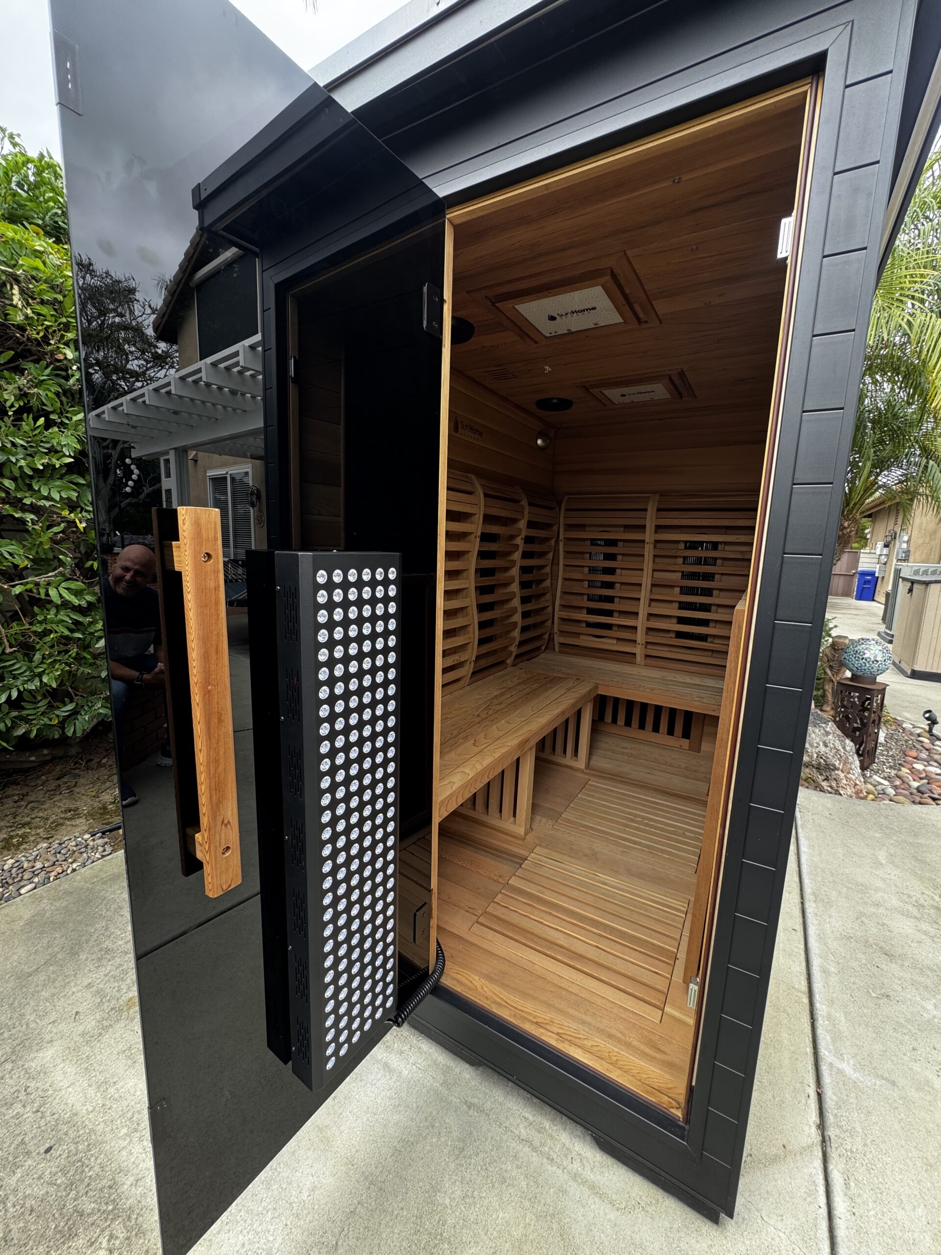 A modern outdoor infrared sauna with a glass door, wooden interior benches, and infrared panels, situated on a concrete patio near plants and trees.