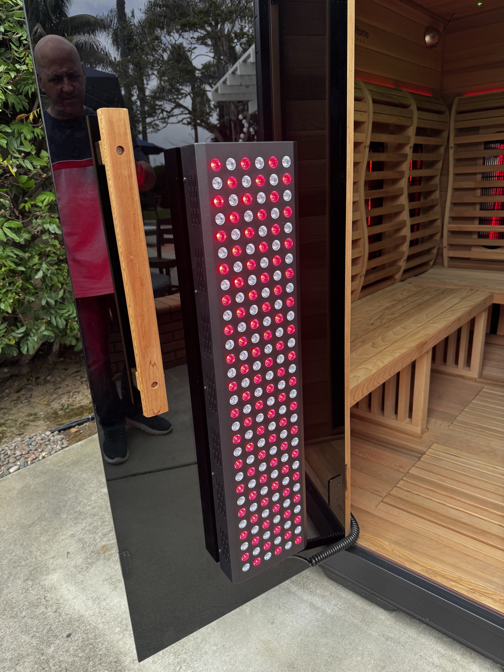 A red light therapy panel is mounted on the door of a wooden sauna with benches inside; a person stands outside the open door.