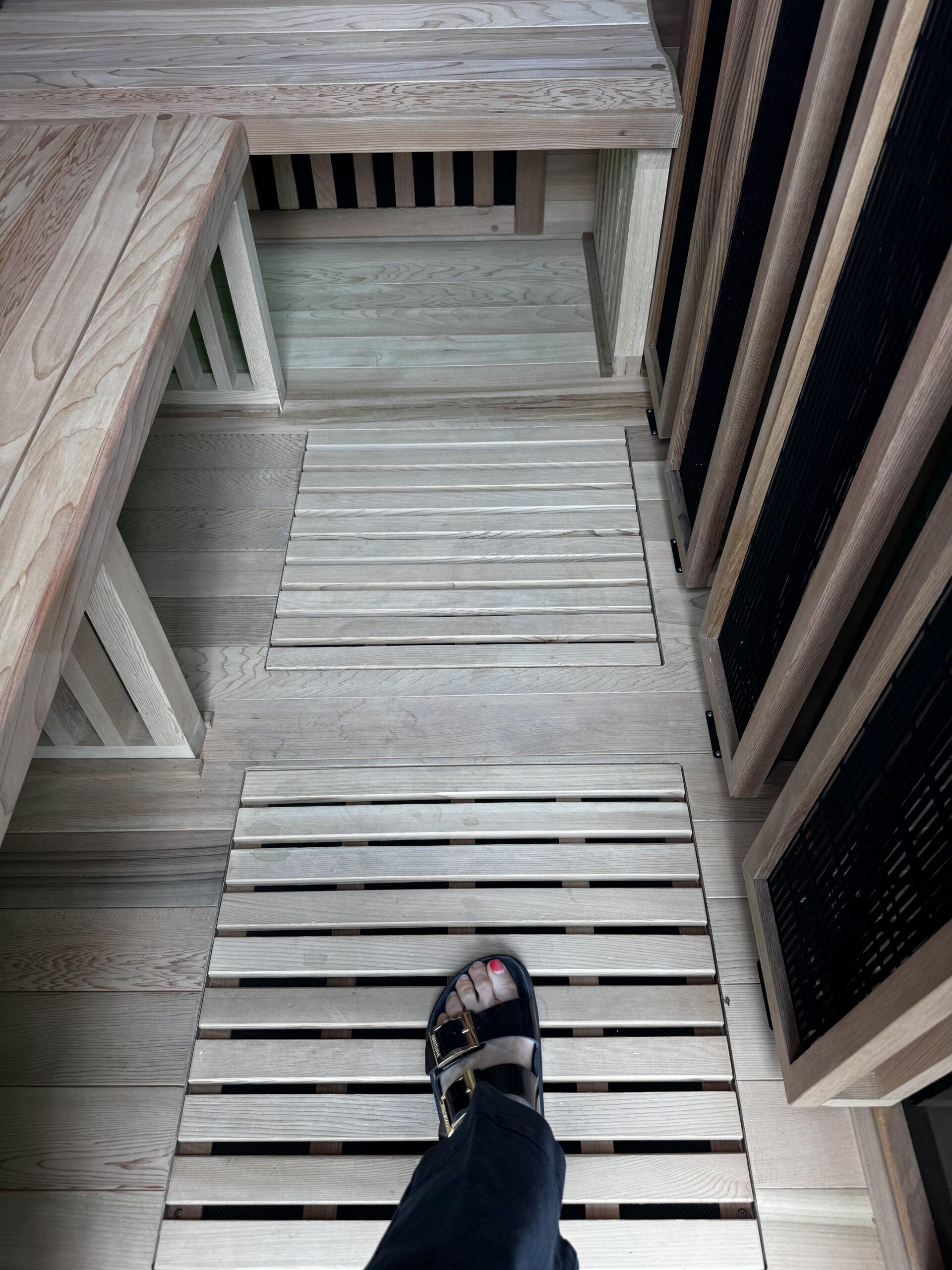 A person wearing black sandals stands on a wooden slatted floor inside a light-colored wooden sauna or steam room.