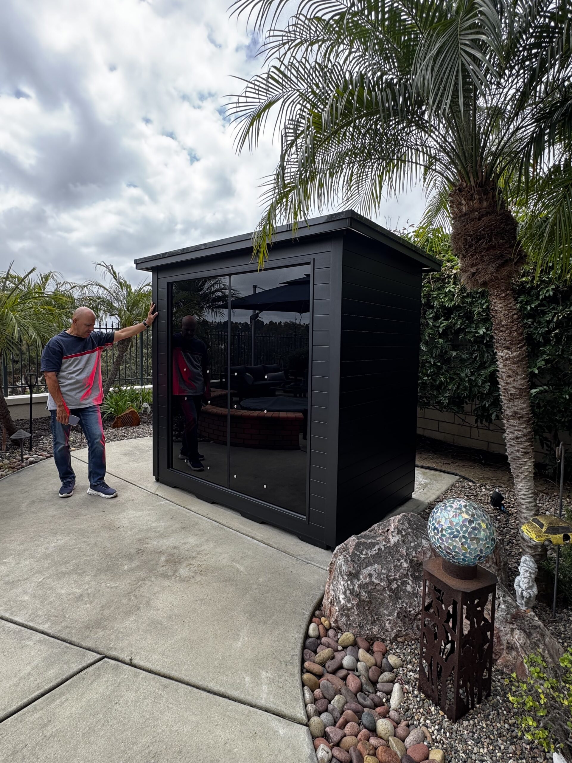 A man stands next to a small black outdoor shed with glass doors, surrounded by palm trees, rocks, and garden decorations on a concrete patio.