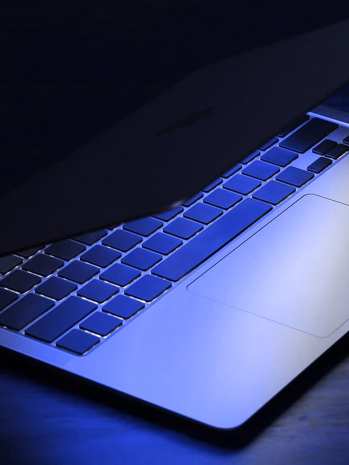 A partially closed laptop with a backlit keyboard is illuminated by blue light in a dark setting.