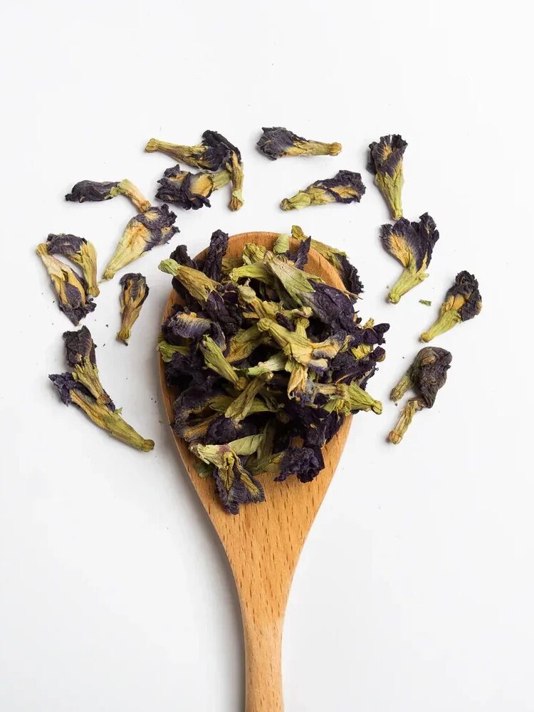 A wooden spoon holds dried purple and green butterfly pea flowers, with additional flowers scattered on a white background.