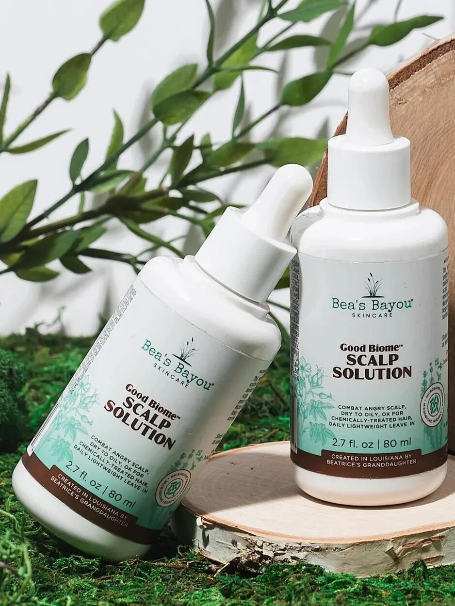 Two bottles of Bea's Bayou Skincare Good Biome Scalp Solution are displayed on a mossy surface with green leaves in the background.