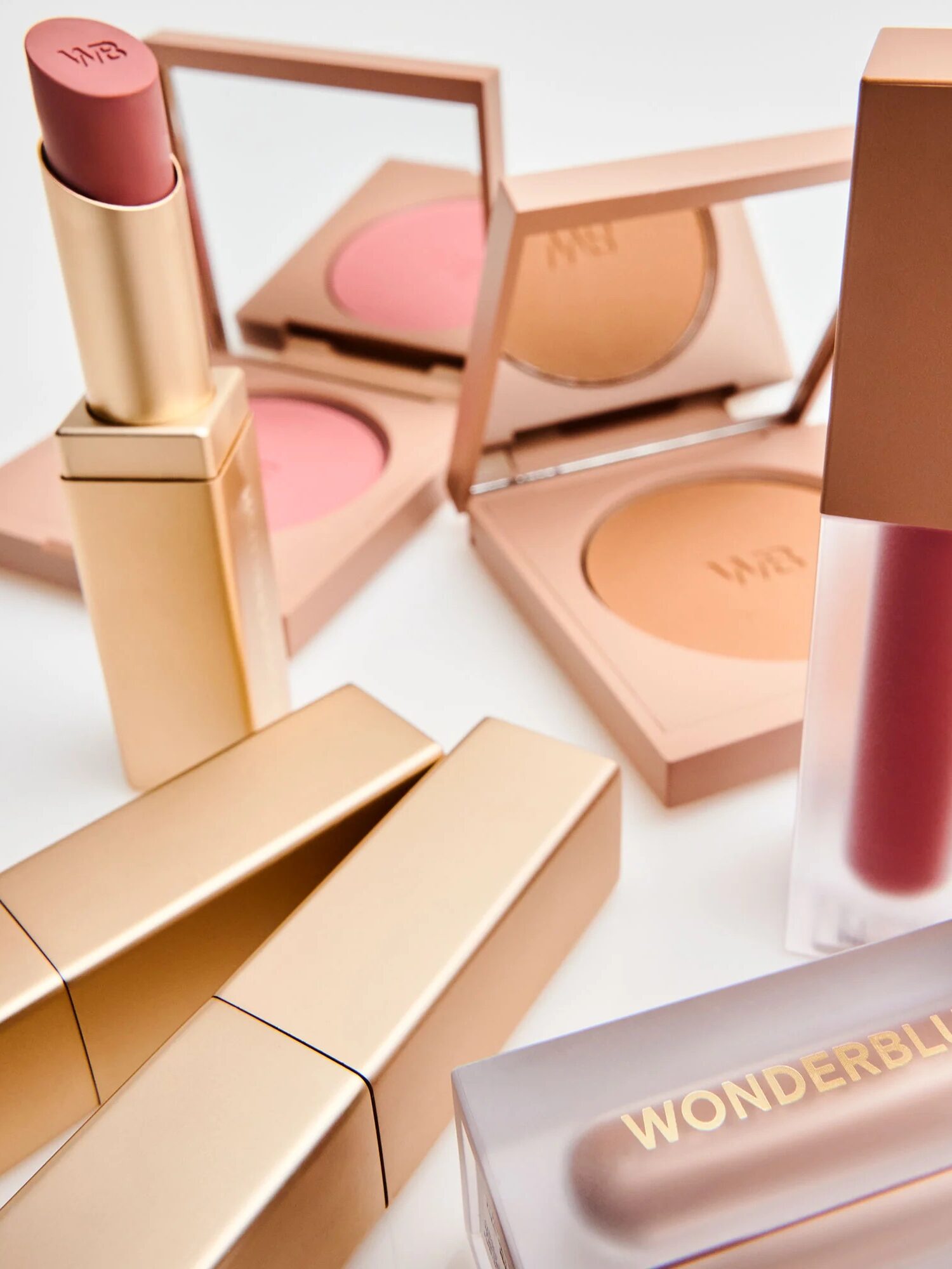 Assorted makeup products, including lipstick, blush compacts, and a liquid product, in gold and frosted packaging arranged on a white surface with a mirror in the background.