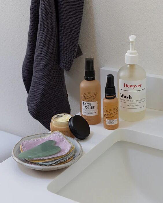 A bathroom countertop with skincare products, a stack of reusable cotton pads, a gua sha tool, and a dark towel hanging on the wall.