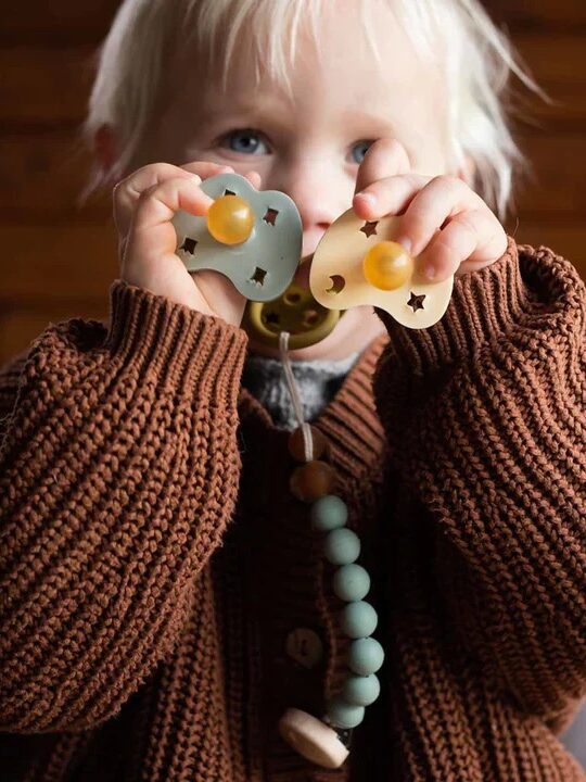 A young child in a brown sweater holds two pacifiers up to their face while sucking on a third pacifier.