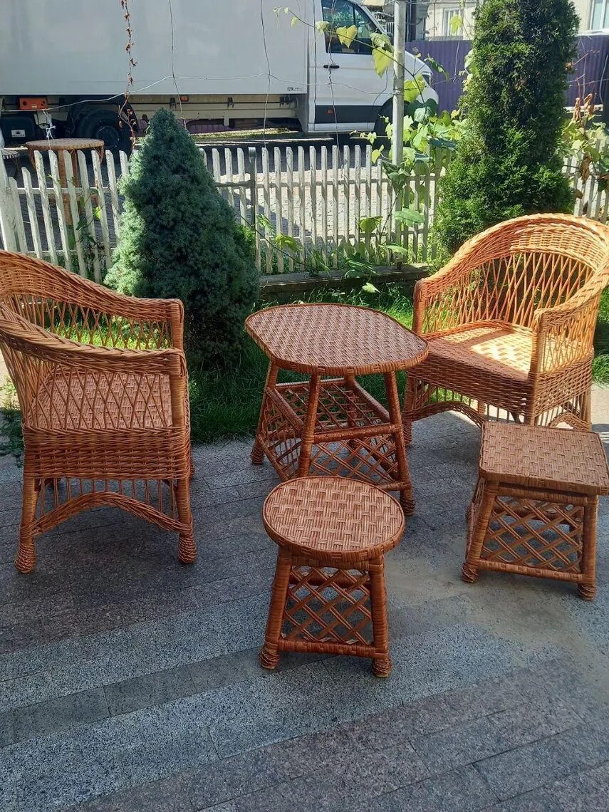 A set of four wicker furniture pieces, including two chairs, one table, and one stool, arranged on a patio with a fence, plants, and a truck in the background.