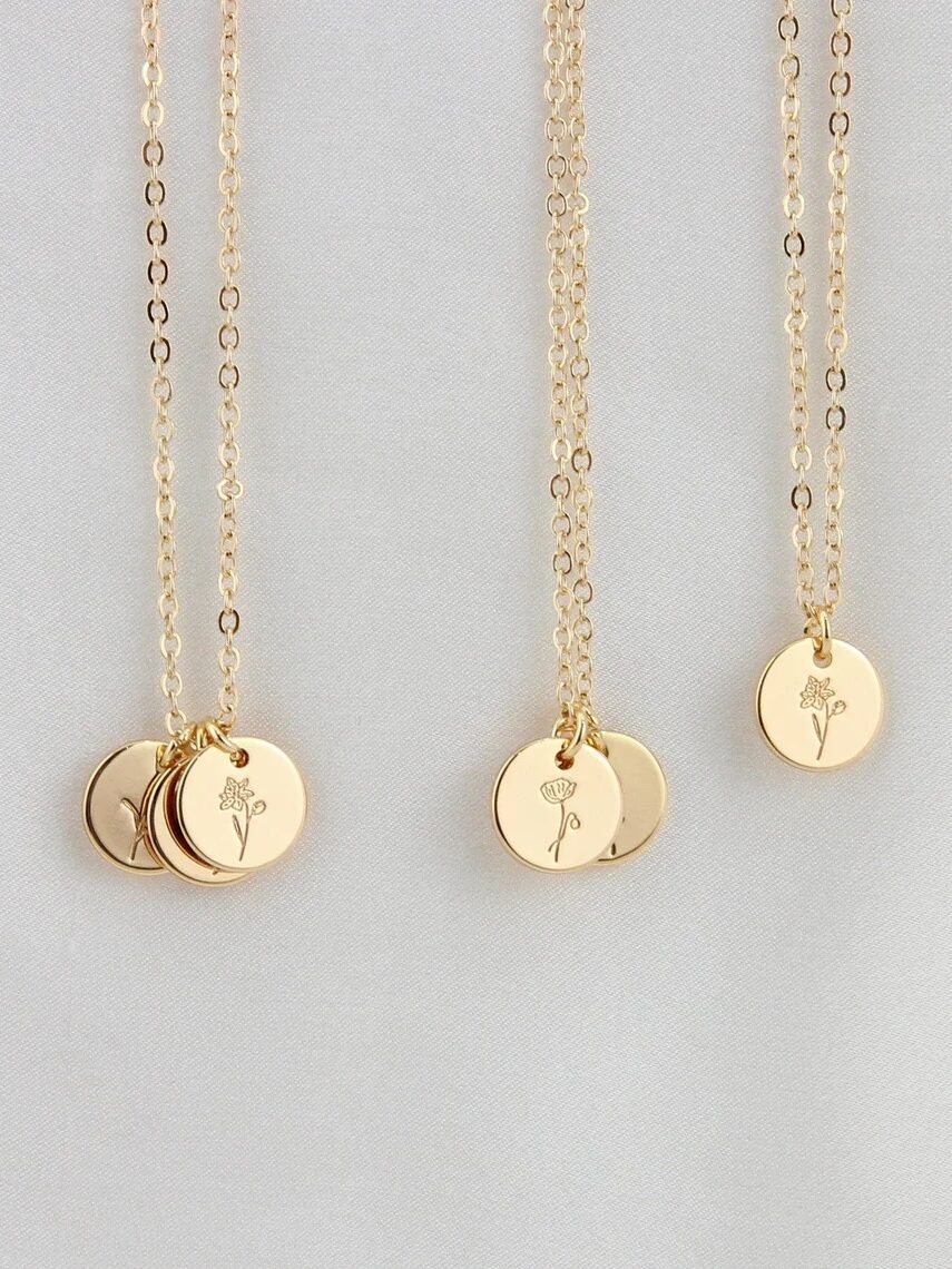 Three gold necklaces with round pendants, each engraved with a different flower design, displayed on a light, neutral background.