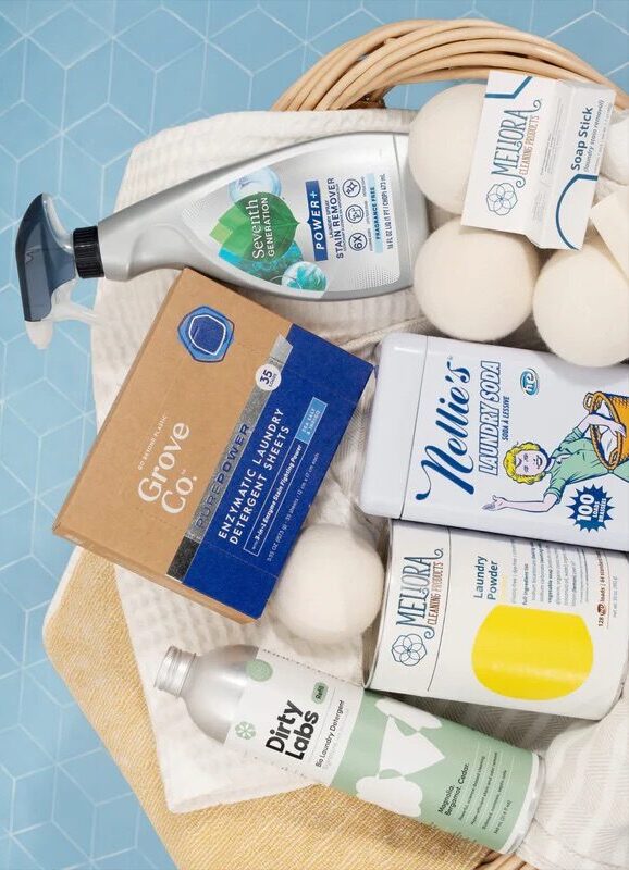 A basket contains eco-friendly laundry products, including detergent sheets, stain remover spray, laundry soda, laundry powder, wool dryer balls, and a soap stick.