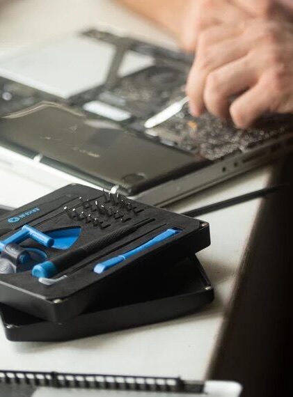 A person repairs an open laptop using a precision screwdriver set, with various tools and components visible on a white desk.