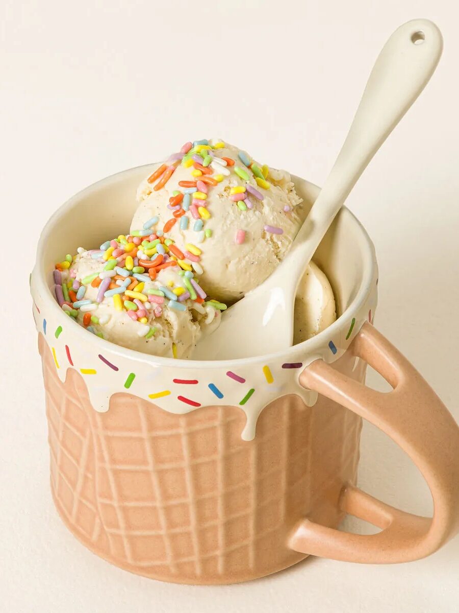A ceramic mug designed to look like a waffle cone holds two scoops of vanilla ice cream topped with colorful sprinkles, with a white spoon resting inside.