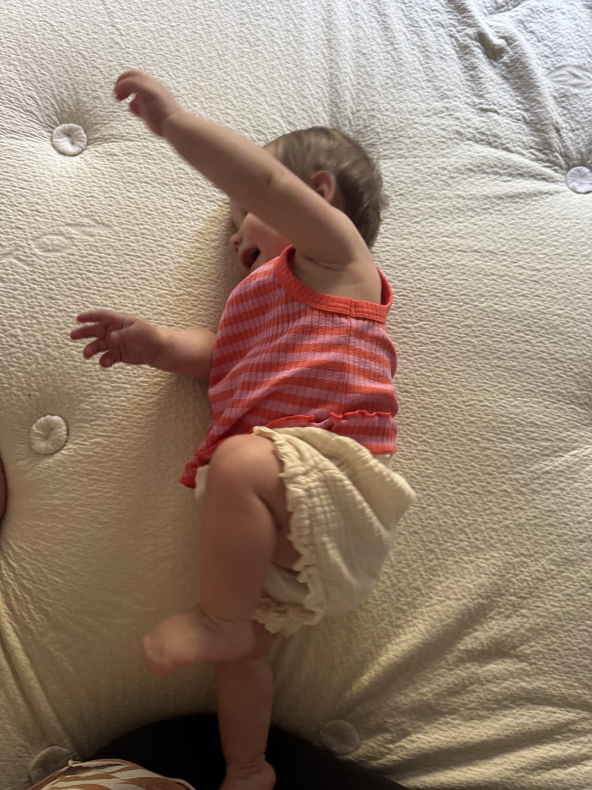 A baby wearing a red checkered top and cream-colored shorts lies on a textured light-colored surface with one arm raised.