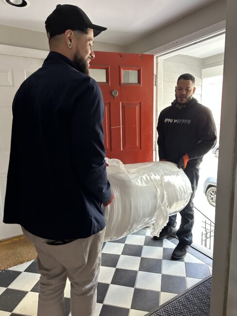 Two men carry a large, wrapped item through the doorway of a house with a red door and black-and-white checkered floor.