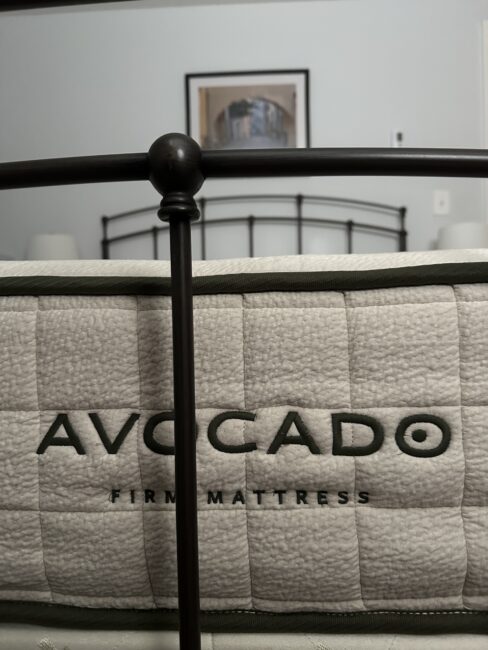 Close-up of an Avocado brand firm mattress, with a metal bed frame in the foreground and a framed picture on the wall in the background.
