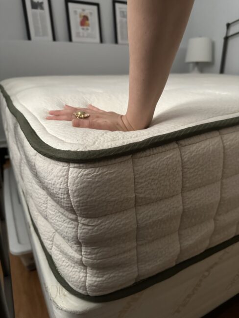 A hand presses down on the surface of a thick, white mattress with a green trim, showing its softness. Framed pictures and a lamp are visible in the background.