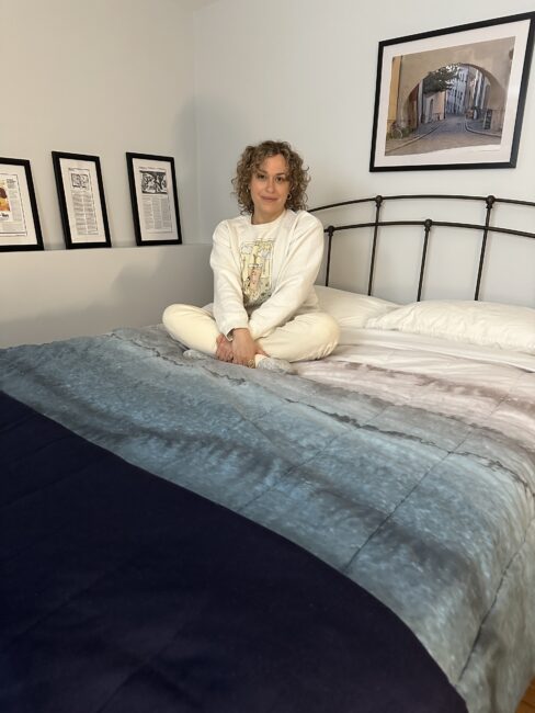 A person with curly hair sits cross-legged on a bed in a neatly decorated bedroom with framed art on the wall.