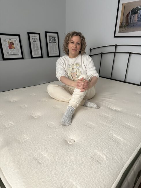 A person sits cross-legged on an unmade mattress in a bedroom with framed pictures and articles on the walls.