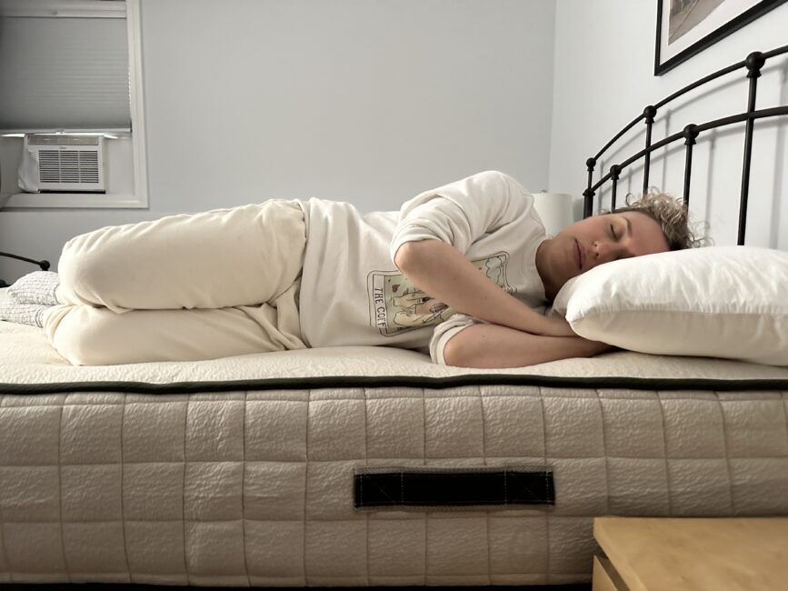 A person lies on their side on a bed, eyes closed, resting their head on a white pillow in a simply furnished bedroom.