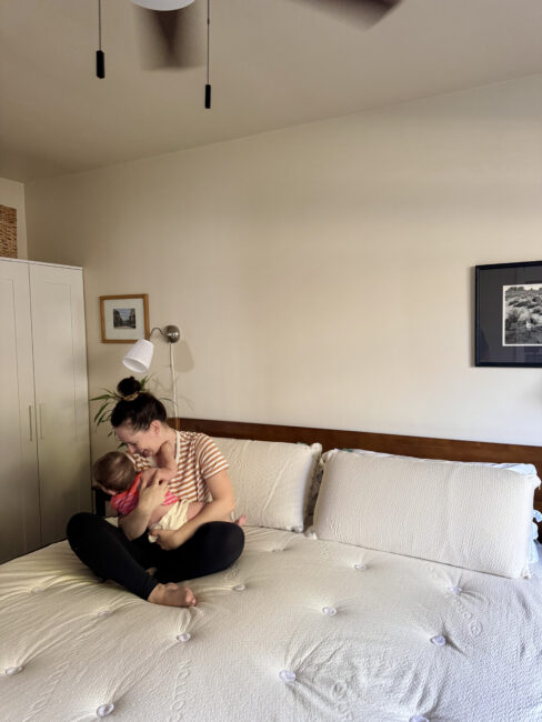 A woman sits cross-legged on a white bed, holding a small child in her lap in a simply decorated bedroom with neutral tones.