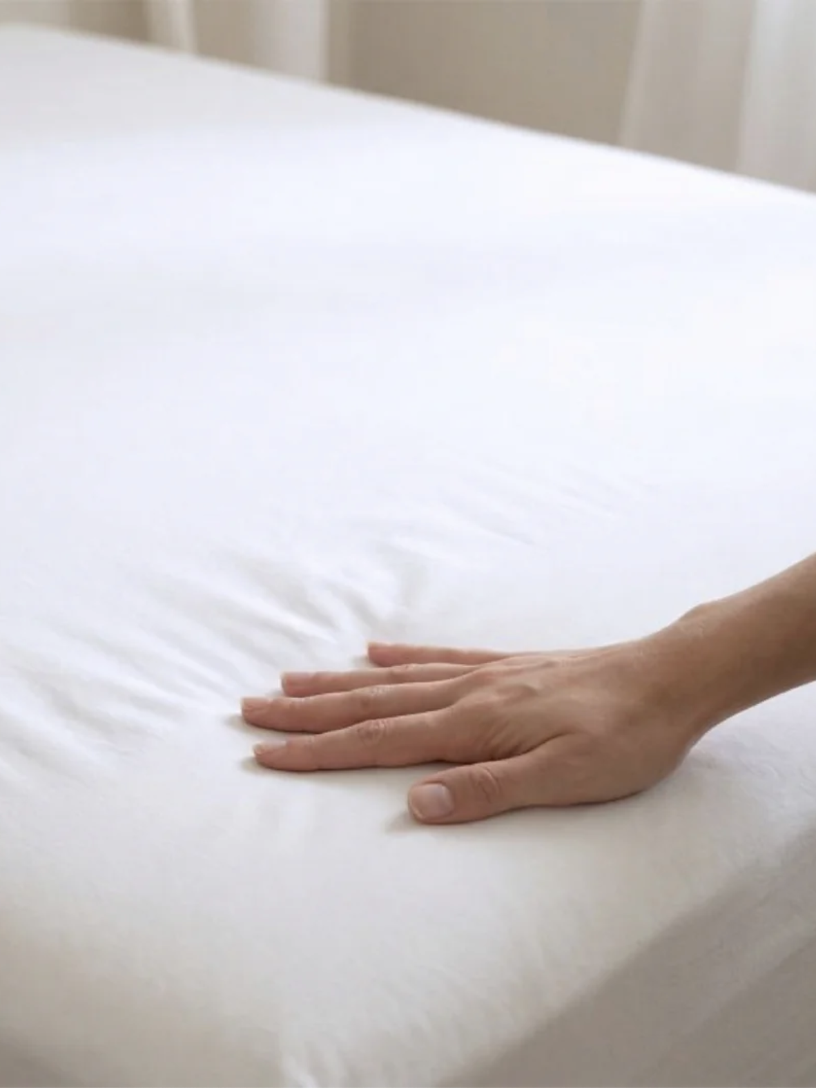 A hand pressing down on a neatly made bed with a white fitted sheet.