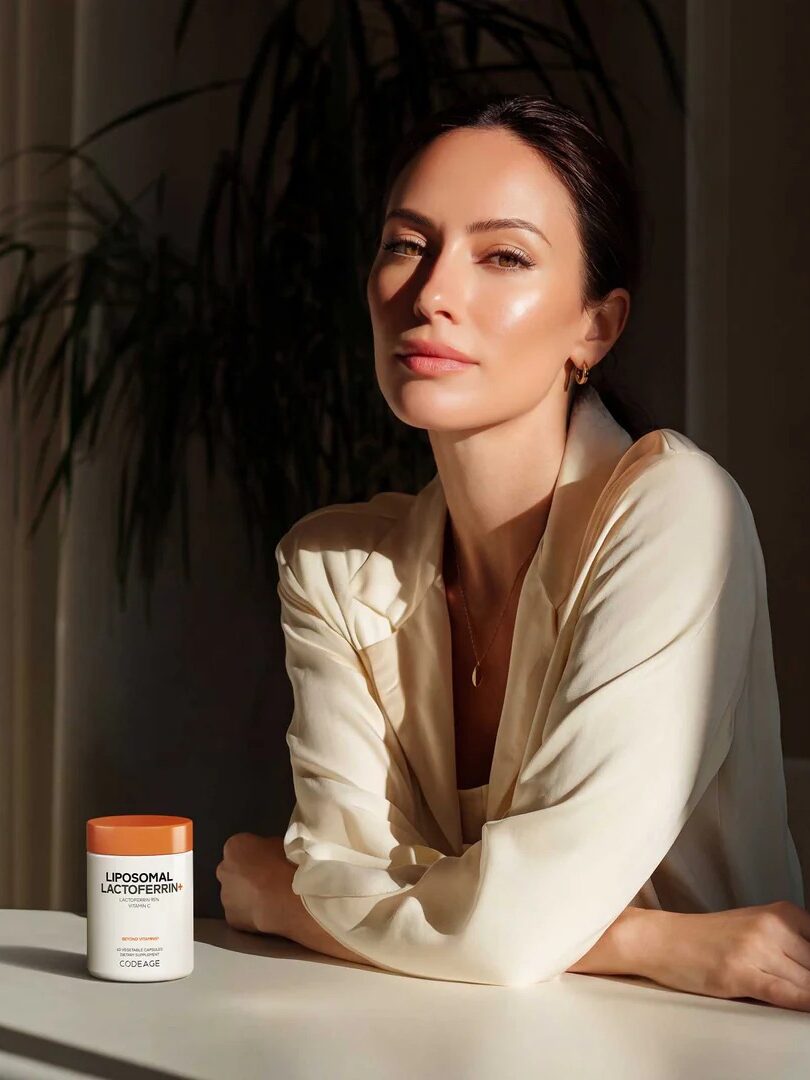 A woman in a cream blouse sits at a table with a container labeled "Liposomal Lactoferrin" in front of her, with sunlight casting shadows in the background.