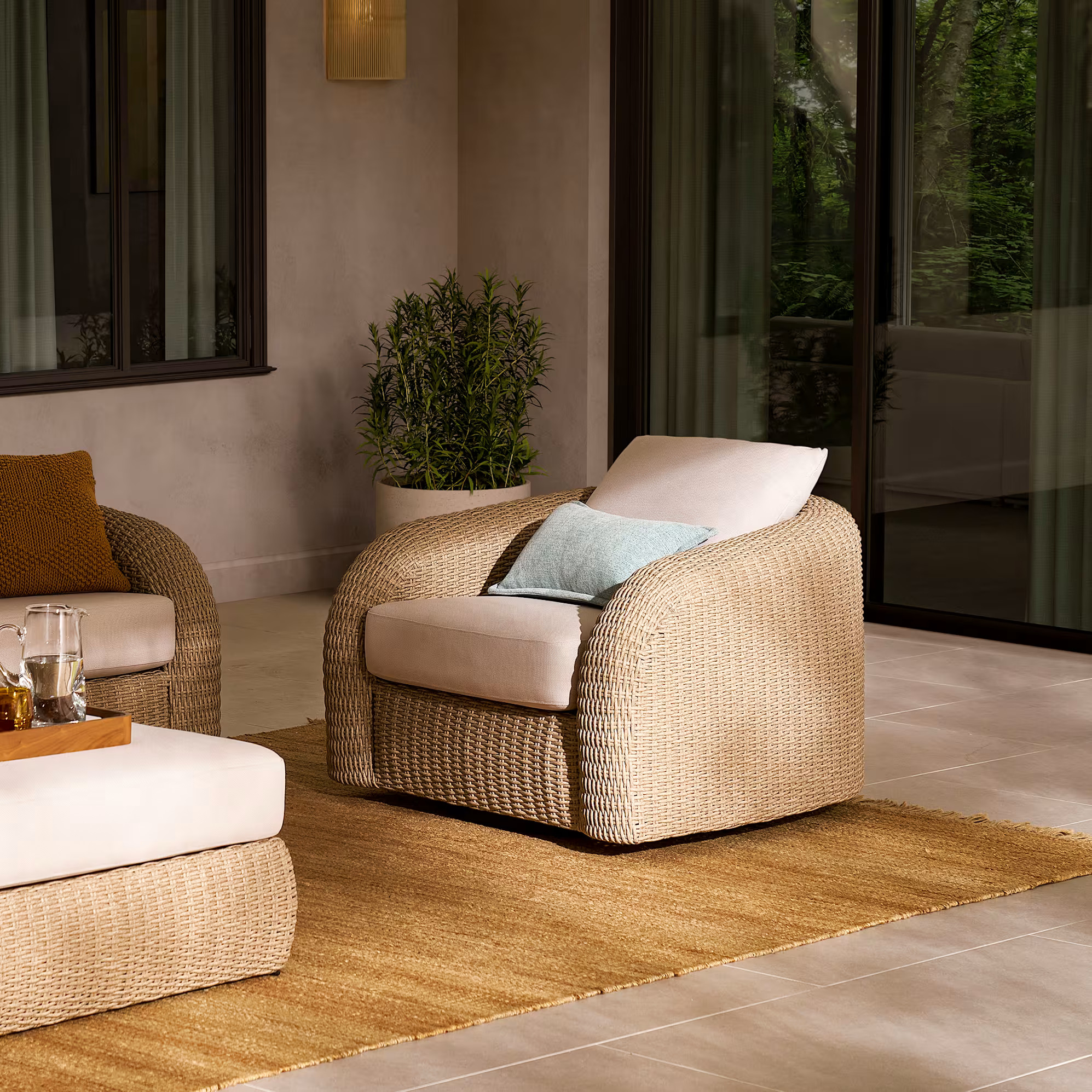 A wicker armchair with cream cushions and a light blue pillow sits on a woven rug in a modern indoor-outdoor space near a glass door and a potted plant.