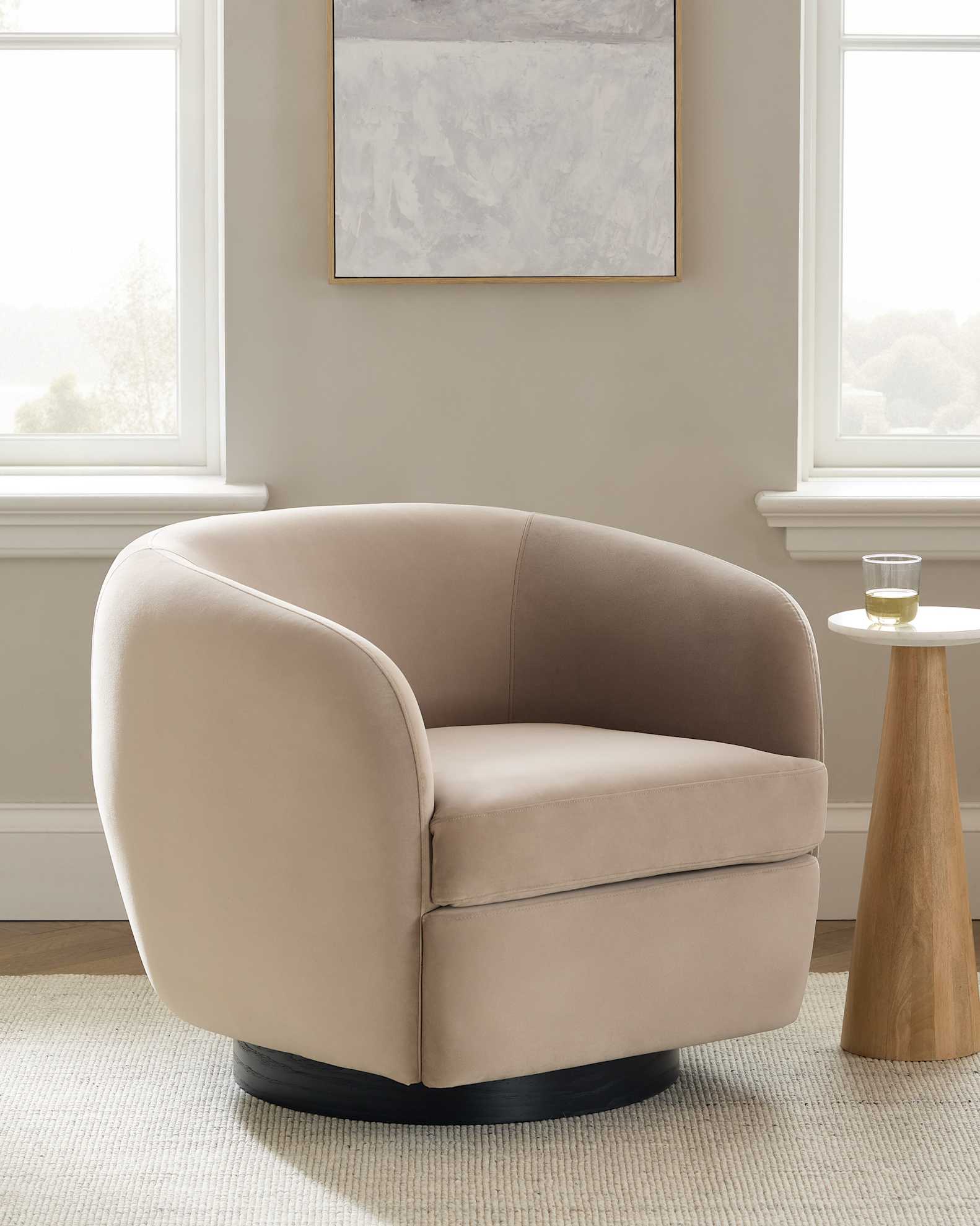 A beige, modern swivel chair with curved arms sits next to a small round side table in a bright room with two windows and a framed wall art.