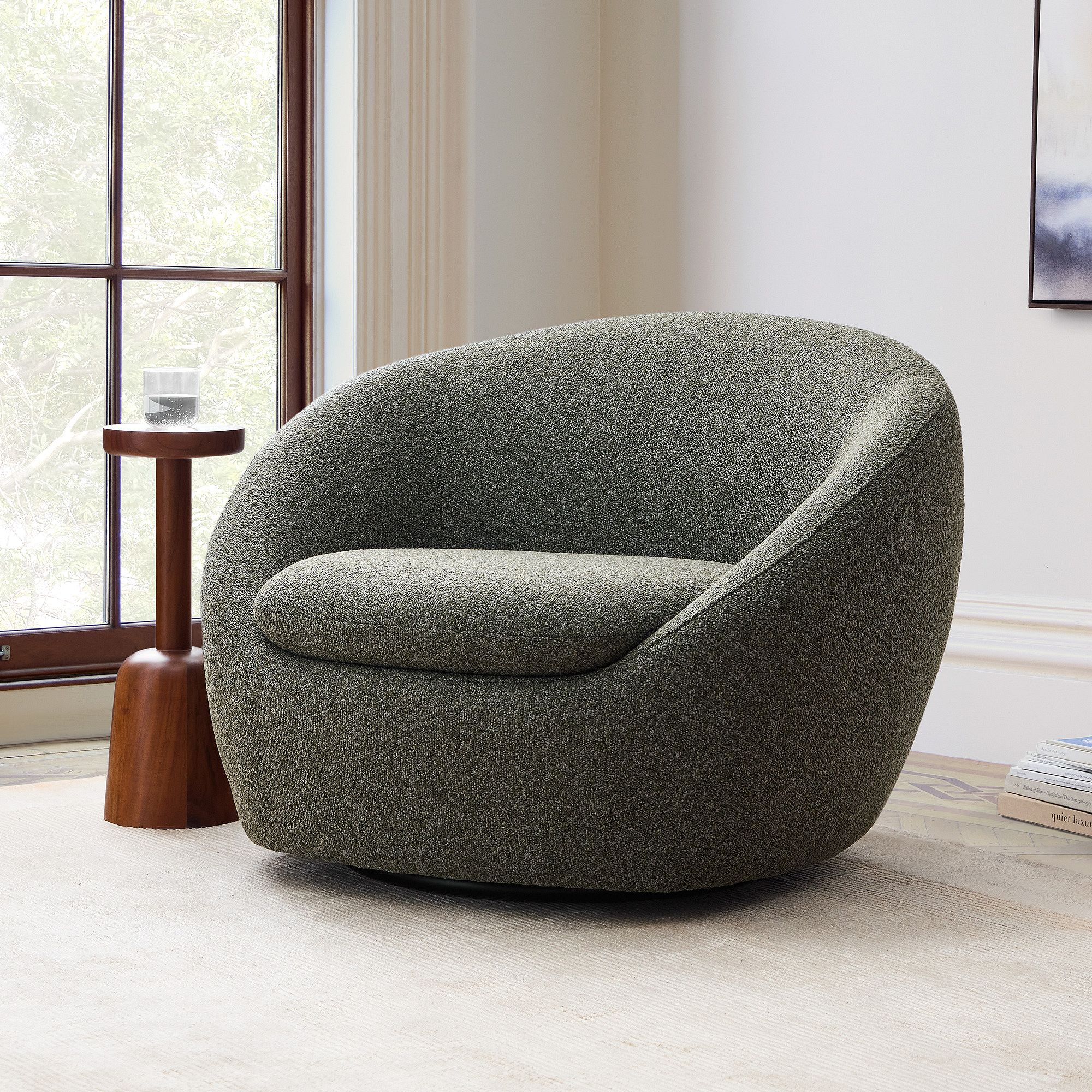A modern, rounded gray armchair sits on a light rug next to a small wooden side table with a glass, in a bright room with large windows.