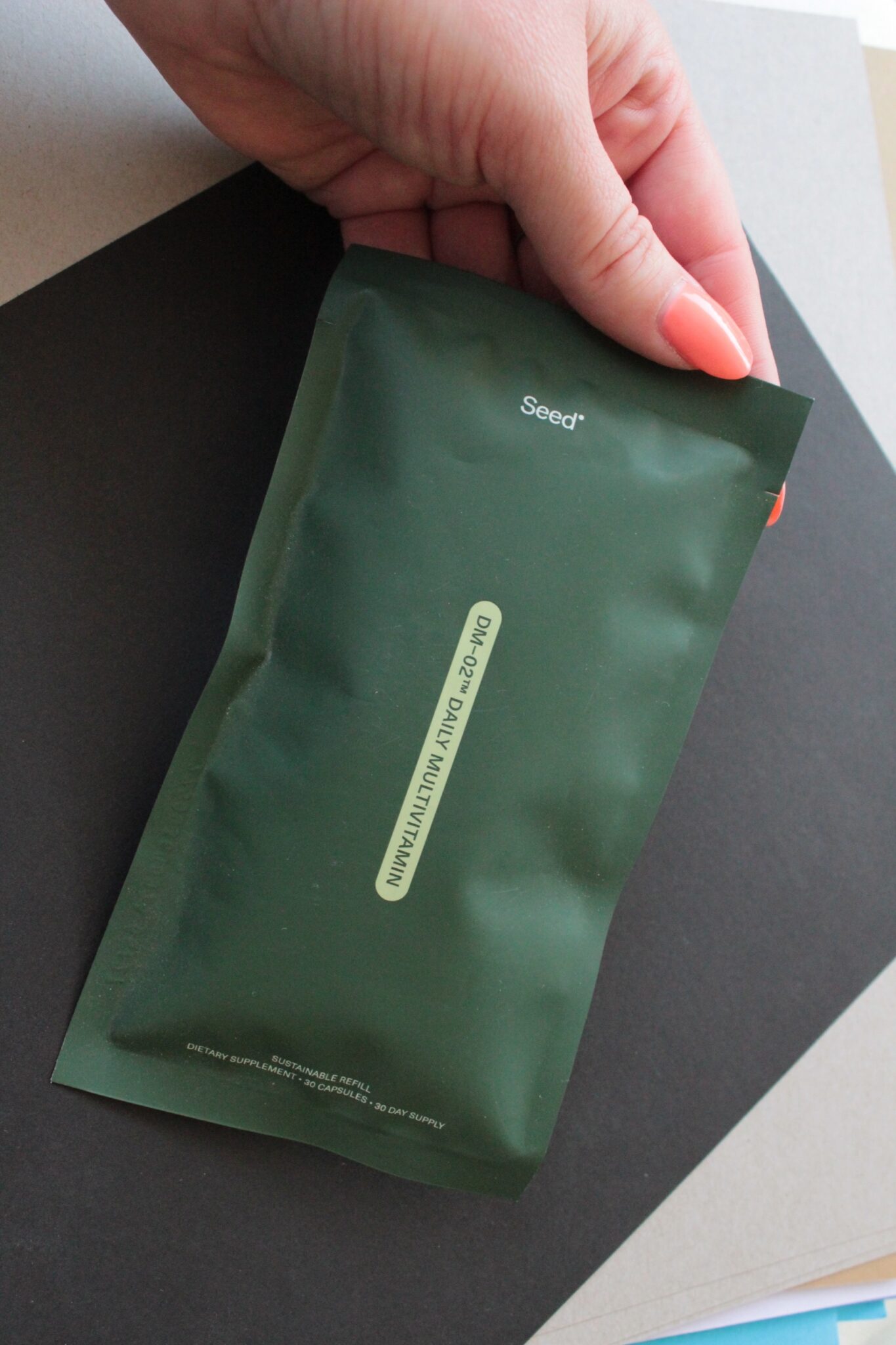 A hand holds a dark green Seed-branded biodegradable refill packet against a background of layered papers.
