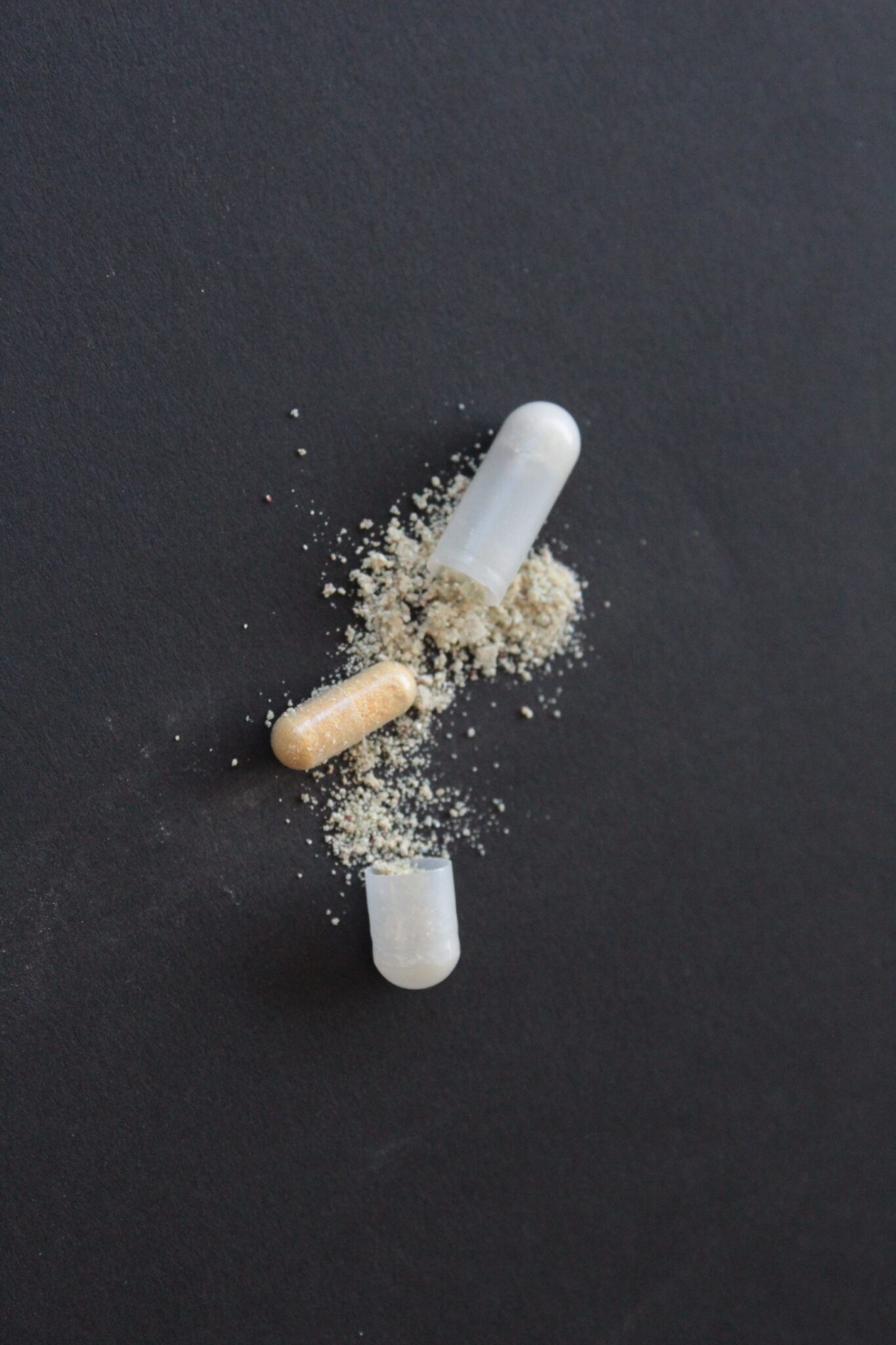 Two capsules, one clear and one beige, with powder spilling out onto a black surface.