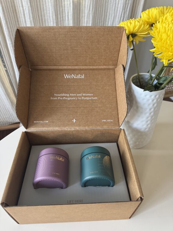 An open WeNatal box with two supplement containers, one purple and one teal, on a table next to a white vase with yellow flowers.